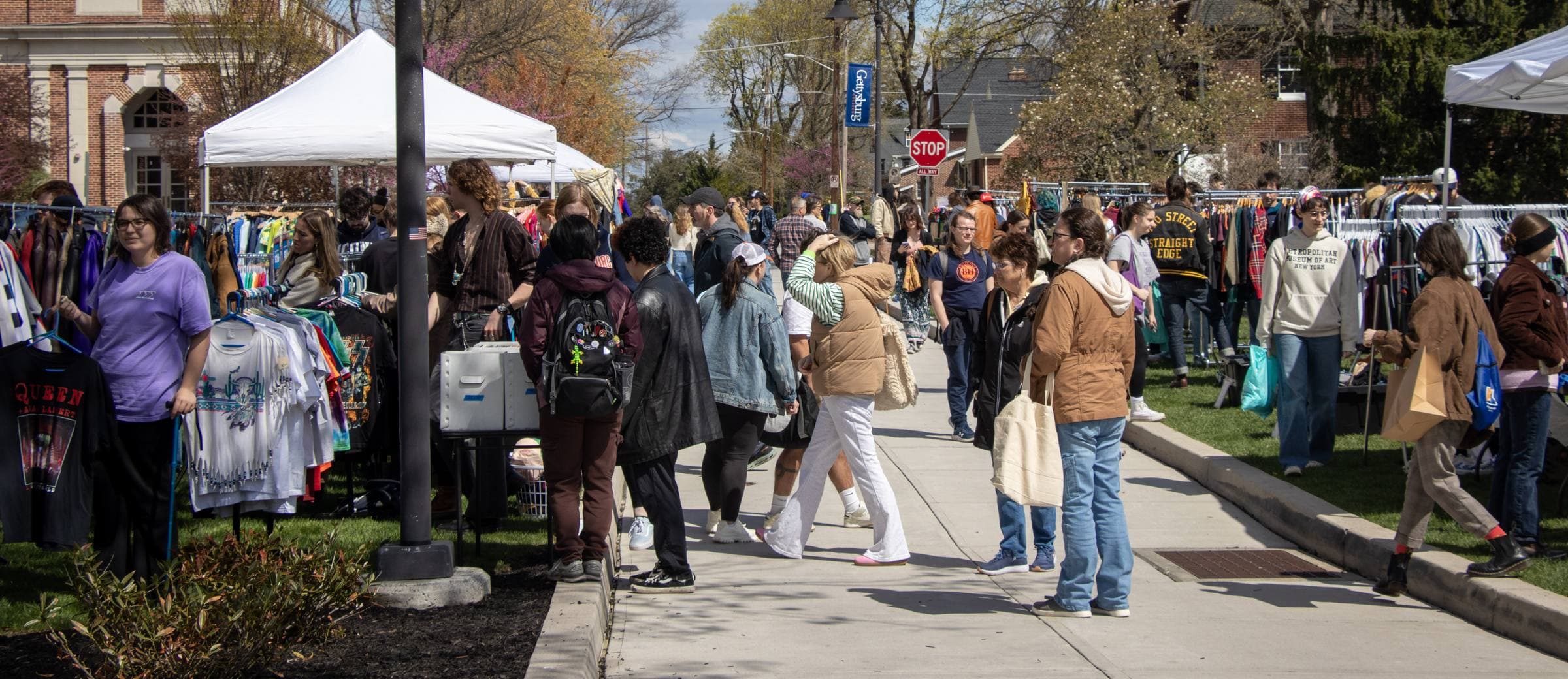 Shoppers browsing racks of vintage clothing between white canopy tents at a spring street market.